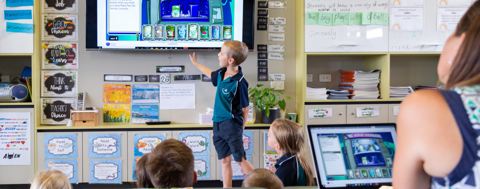Primary school student standing at a wall-mounted display in the front of a class, pointing to what's on the screen.