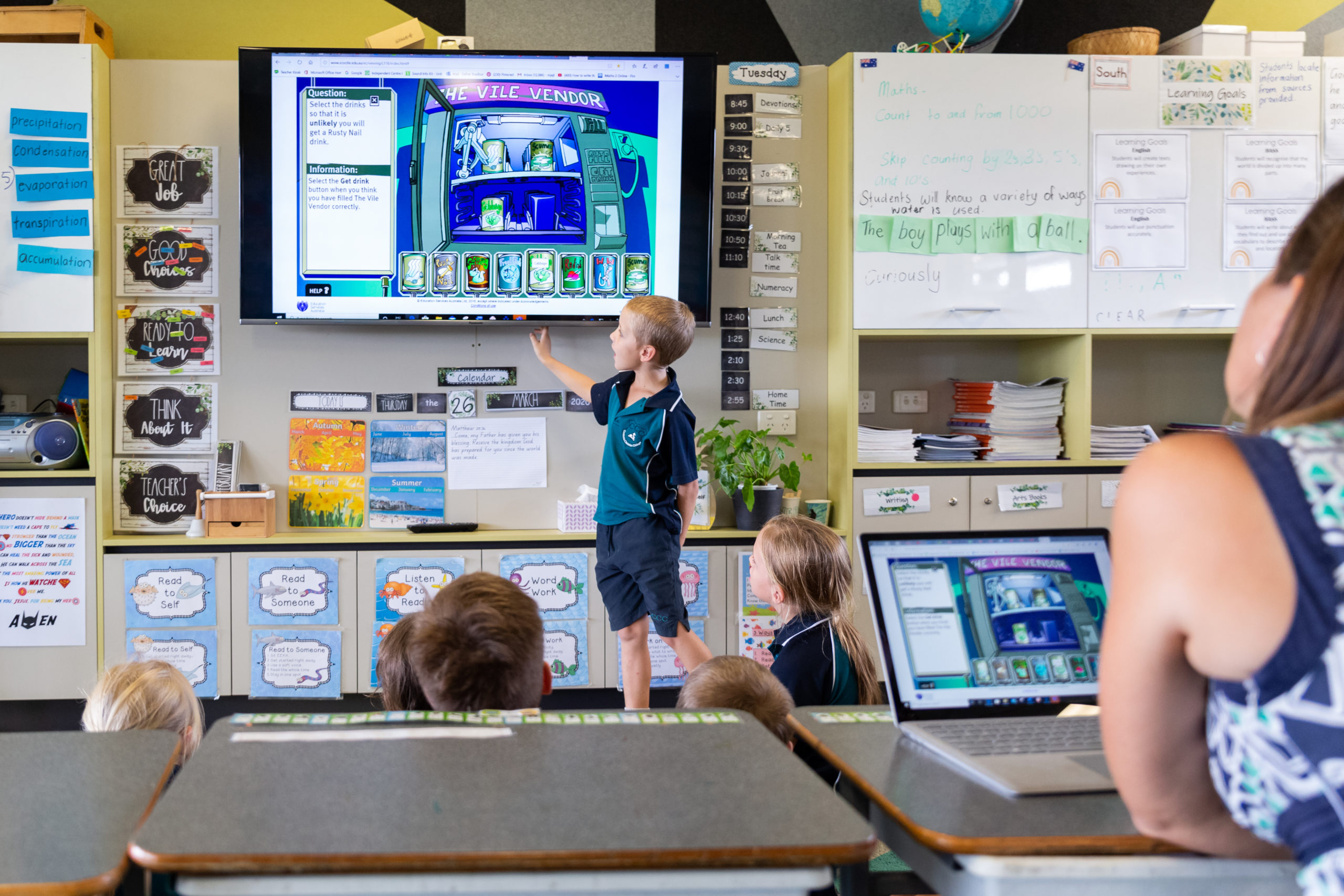 Primary school student standing at a wall-mounted display in the front of a class, pointing to what's on the screen.