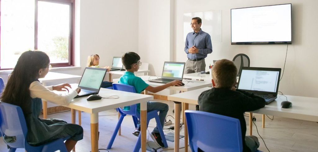 A classroom with four people using laptops, and a presentation being displayed on a screen.