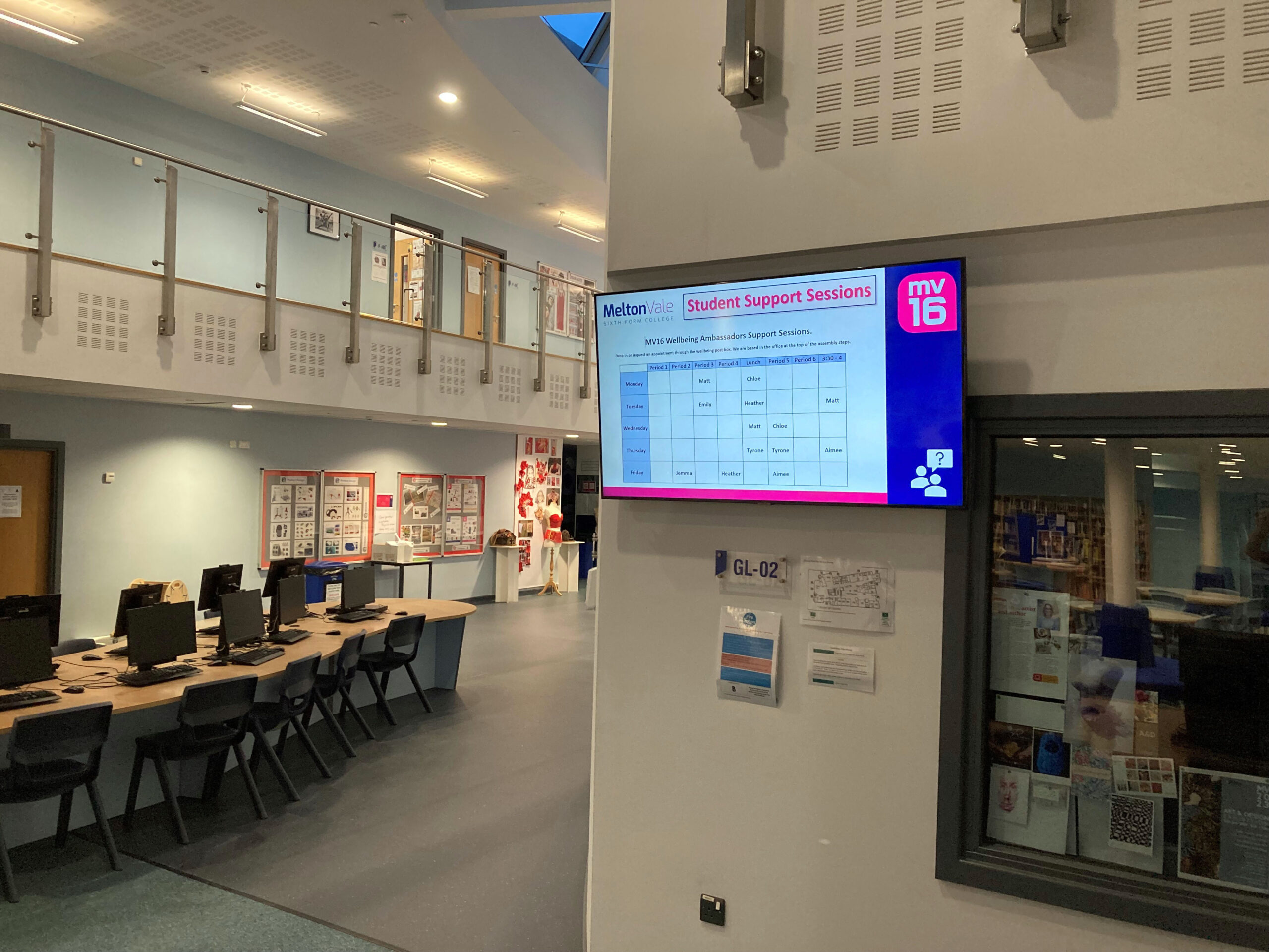 A school hallway with a digital display showing a school schedule hanging from the wall.