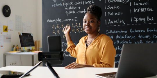 teacher leading her blog online African American teacher leading her blog online about IT while sitting at table in front of smartphone on tripod
