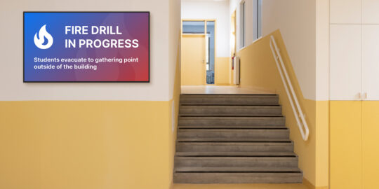 An empty school hallway with a digital sign displaying an emergency message explaining that a fire drill is in progress.