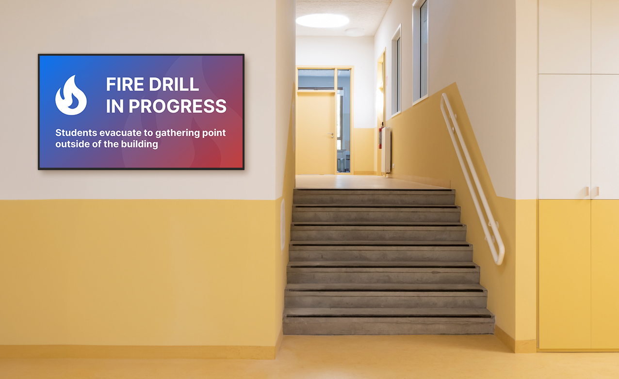 An empty school hallway with a digital sign displaying an emergency message explaining that a fire drill is in progress.