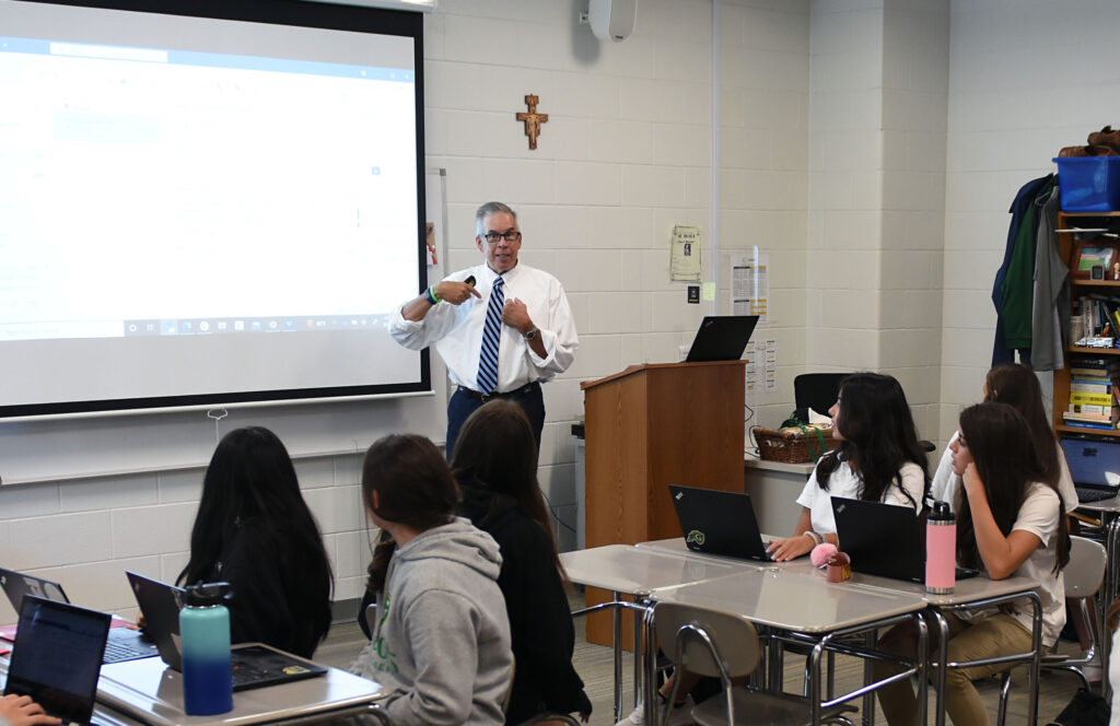 High school classroom with a male teacher sharing a lesson with a group of students. He is standing at a projector screen, holding a remote, and sharing content with the class.