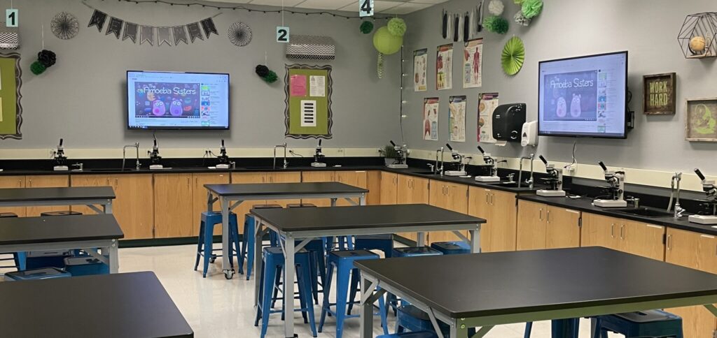 Science lab classroom with black countertops, stools, microscopes, and a TV displaying educational material on the far wall.
