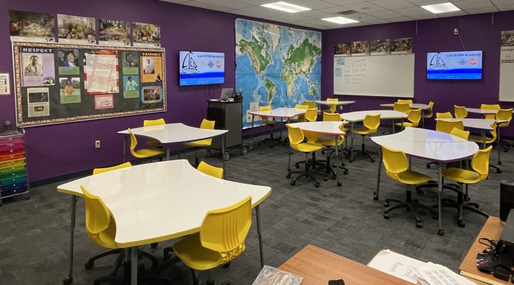 A vibrant classroom with purple walls, yellow chairs around white tables, educational posters, and a large world map.