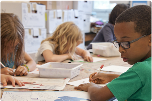 Elementary school-age boy wearing glasses writes in his workbook at a table in a classroom alongside classmates.