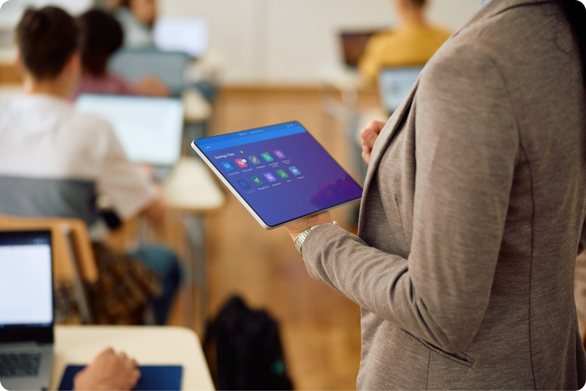 A person in a grey blazer holding a tablet with various app icons, standing in a room with others who are using laptops, indicating a collaborative classroom environment.