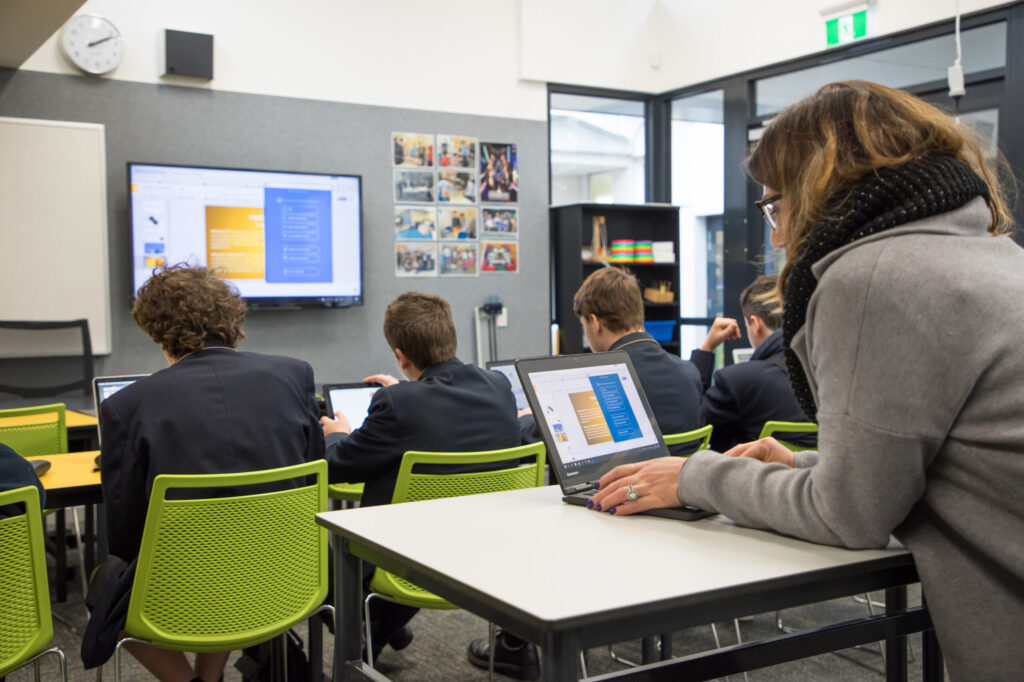 Students and a teacher in a modern classroom, working on laptops and focusing on a presentation displayed on a screen.