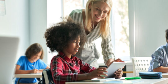 Happy mature female teacher educator helping African American junior school kid girl student using digital tablet computer education program app technology during elementary class lesson in classroom.