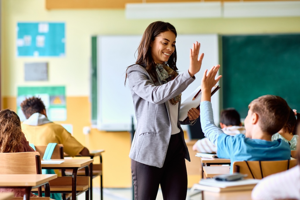 Why Teacher Wellbeing Drives Student Success: Tips and Strategies 1 Happy elementary school teacher gives high-five to her student during class in classroom.