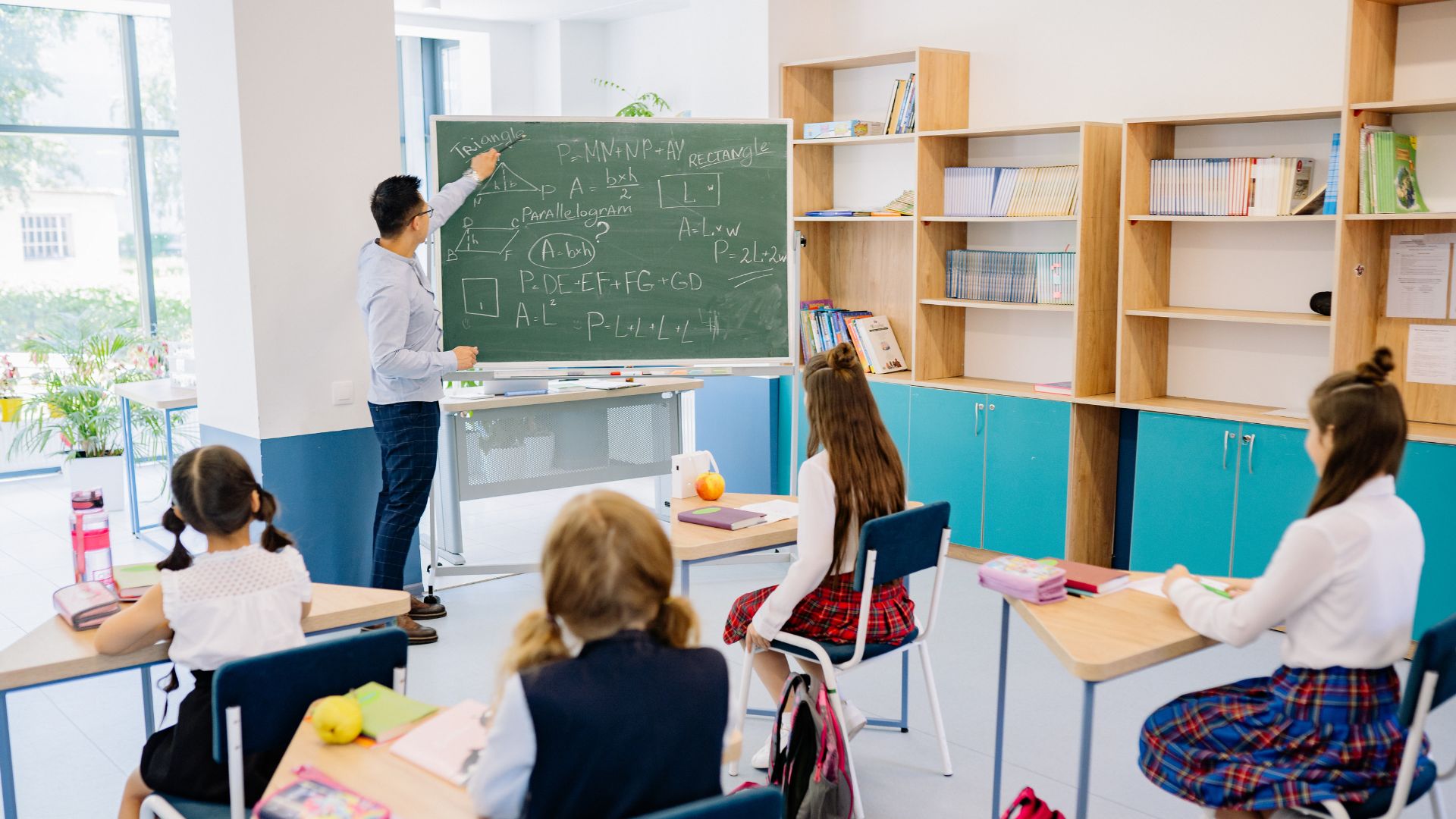 A male classroom teacher writing math equations on a green chalkboard while students sit at desks in a bright, modern classroom.